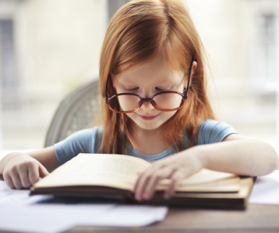 A young girl with red hair and oversized glasses looks down at an open book with a smile.